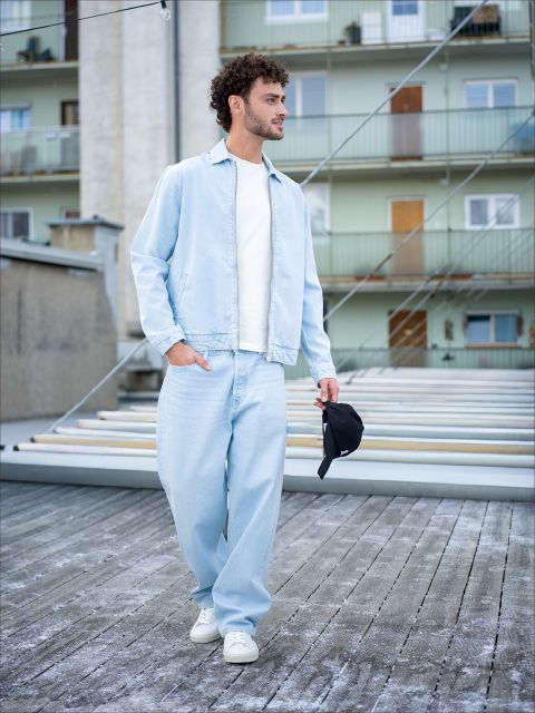 Un hombre con vaqueros azul claro, chaqueta a juego y zapatillas blancas camina al aire libre y sostiene un gorro negro en la mano, con edificios residenciales al fondo.