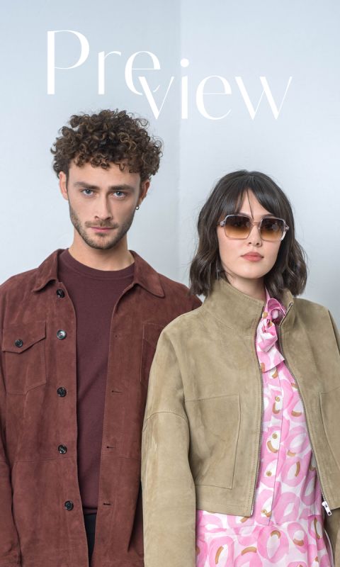 Un hombre con una chaqueta marrón y una mujer con gafas de sol y una chaqueta marrón claro sobre un vestido con estampado rosa están de pie uno al lado del otro frente a un fondo claro y sencillo.