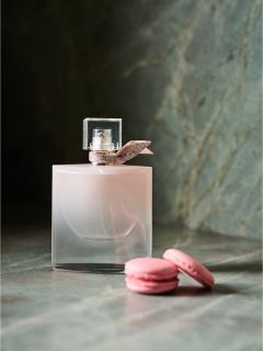 Un frasco de perfume rosa con un lazo está al lado de dos macarons rosas sobre una superficie de mármol.