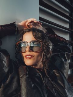 Una mujer con gafas de sol grandes y una chaqueta de cuero negra se reclina con las manos sobre la cabeza, la luz del sol se refleja en sus gafas.