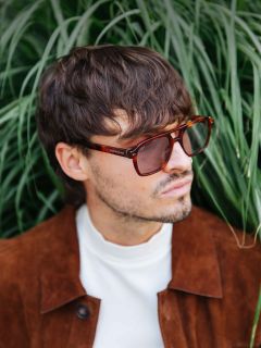 Hombre con cabello castaño despeinado y gafas de sol, con una chaqueta de ante marrón y un jersey de cuello alto blanco, de pie frente a una hierba verde alta.