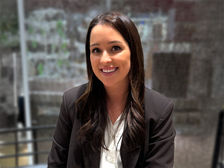 Una mujer con cabello largo y oscuro con un blazer negro y una camisa blanca sonríe mientras está sentada en una casa, con un fondo borroso.