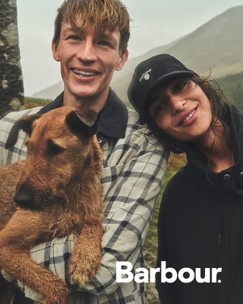 Un hombre sonriente con un perro marrón está de pie junto a una mujer sonriente con un gorro, ambos al aire libre con colinas al fondo. El logotipo de Barbour es visible.