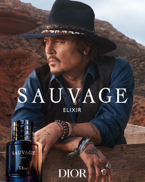 Un hombre con sombrero y chaleco se apoya en una barandilla de madera en el desierto, en primer plano un frasco del perfume Sauvage Elixir Dior con logotipo.
