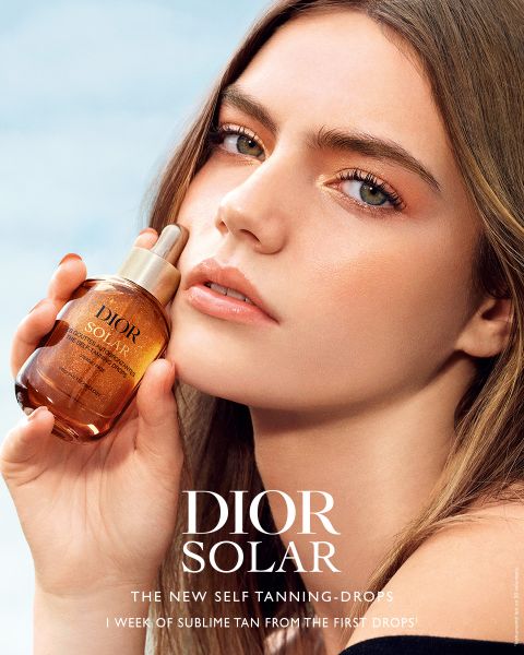 Una mujer con una piel radiante sostiene un frasco de gotas autobronceadoras de Dior Solar cerca de su rostro, donde se puede leer el nombre del producto y el eslogan en letras blancas.