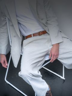 Una persona con un traje gris claro y una camisa blanca está sentada en una silla blanca, se ve un cinturón marrón y su rostro no se ve.