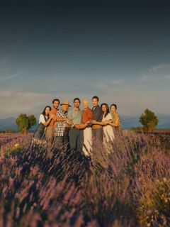 Un grupo de ocho personas está de pie, tomados de la mano, en un campo de lavanda bajo un cielo despejado, rodeados de flores púrpuras en flor.