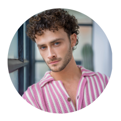 Hombre con cabello castaño rizado y ojos azules, que lleva una camisa de rayas rosas y blancas y mira a la cámara en un interior junto a una ventana.