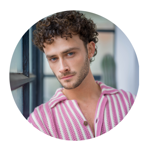 Hombre con cabello castaño rizado y ojos azules, que lleva una camisa de rayas rosas y blancas y mira a la cámara en un interior junto a una ventana.