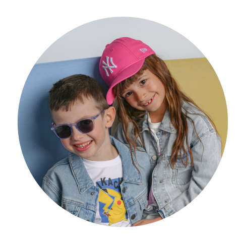 Dos niños sonrientes con chaquetas vaqueras; el niño lleva gafas de sol, la niña una gorra de béisbol rosa y apoya la cabeza en la de él.