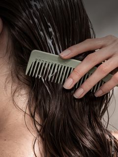 Una persona peina cabello oscuro y húmedo con un peine de dientes anchos, distribuyendo el acondicionador en las hebras.