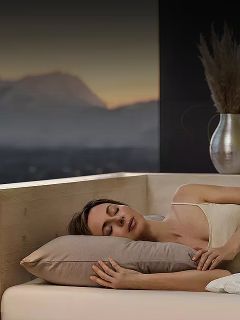Una mujer duerme de lado en la cama con una almohada beige, junto a una ventana que muestra un paisaje montañoso borroso al atardecer.