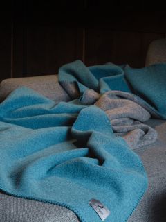 Una manta de lana azul con la parte inferior gris está colocada sobre un sofá gris, con una etiqueta de tela visible en primer plano.