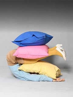 Una persona yace enrollada en el suelo, cubierta por tres grandes cojines coloridos, de los que solo sobresalen sus piernas vestidas de amarillo y zapatos blancos.