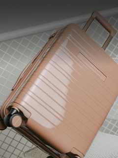 Una maleta rígida brillante de color marrón claro con ruedas y un asa extendida está sobre un suelo de baldosas.