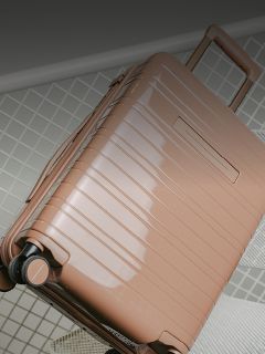 Una maleta rígida brillante de color marrón claro con ruedas y un asa extendida se encuentra sobre una superficie embaldosada.