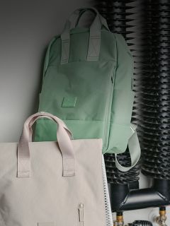 Una mochila verde menta y una bolsa de mano beige cuelgan frente a tubos y carretes negros en una pared.