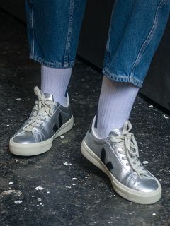 Persona con zapatillas deportivas plateadas metálicas con suelas blancas, calcetines blancos y vaqueros azules de pie sobre un suelo oscuro y texturizado.