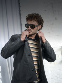 Hombre de pelo rizado con gafas de sol, jersey de rayas y chaqueta de cuero negra ajustándose el cuello frente a una pared de ladrillo blanco.