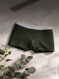 Los bóxers verde oscuro para hombre con cinturilla blanca se exhiben sobre una superficie texturizada, con hojas de eucalipto cerca en una luz suave y natural.