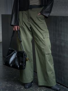 Persona que lleva pantalones cargo verde oliva, zapatos negros y una chaqueta negra, y que sostiene un bolso de mano negro brillante a su lado.