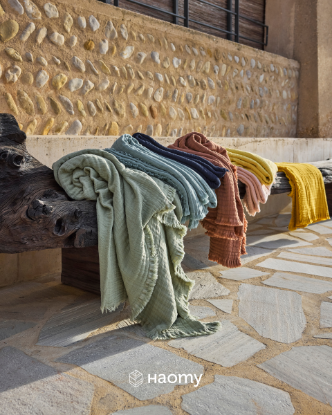 Coloridos y estructurados cobertores colocados sobre una rústica banca de madera al aire libre, con una pared de piedra y una acera al fondo. Logotipo de Naomy en la parte inferior.