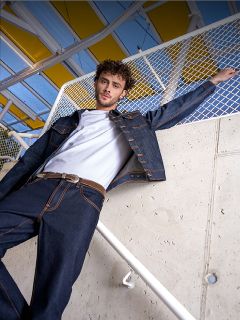 Un hombre con una camiseta blanca, una chaqueta vaquera oscura y vaqueros está de pie en una barandilla apoyada en una pared de hormigón y una valla, con un toldo amarillo encima.