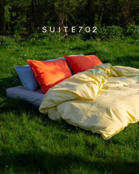 Una cama con almohadas de color naranja brillante y rojo y una colcha amarilla está al aire libre sobre hierba verde a la luz del sol. Encima pone "SUITE702".