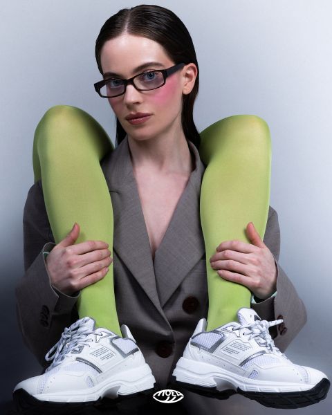 Una mujer con un blazer gris lleva gafas y zapatillas blancas, y sostiene sus piernas sobre los hombros con medias de color verde brillante, delante de un fondo sencillo.