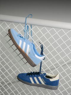 Dos zapatillas Adidas azules con rayas blancas cuelgan de sus cordones en una valla metálica blanca, una azul claro y otra azul oscuro.