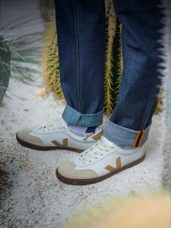 Persona con vaqueros remangados de pie sobre suelo arenoso cerca de cactus, con zapatillas blancas con detalles en beige y marrón y un logotipo en "V" en el lateral.