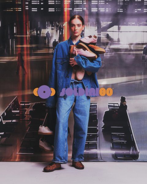 Una mujer con chaqueta vaquera y vaqueros está de pie en una terminal de aeropuerto y sostiene varios pares de zapatillas deportivas en sus brazos.