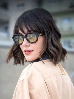 Mujer con cabello oscuro ondulado, con gafas de sol grandes y una chaqueta beige, mirando por encima del hombro al aire libre, con edificios borrosos al fondo.
