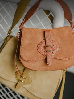 Dos bolsos de mano de ante, uno marrón claro y otro beige, con correas de cuero y cierres dorados, colgados sobre un fondo de rejilla metálica blanca.