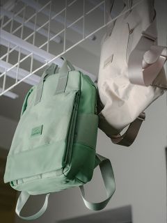 Dos mochilas, una verde menta y otra beige, cuelgan de una estructura metálica blanca en una habitación bien iluminada.