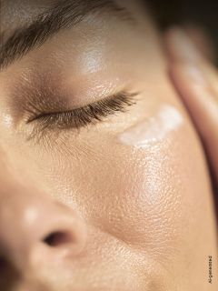 Un primer plano del rostro de una persona con los ojos cerrados, que muestra una piel lisa y radiante y un toque de crema debajo del ojo.