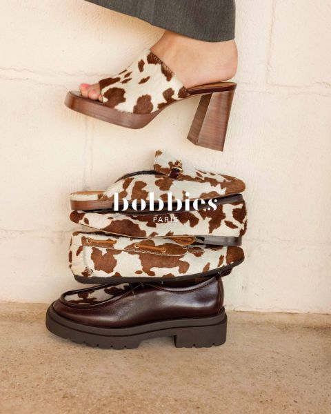 Cuatro zapatos de moda, incluidos tacones con estampado de vaca, mocasines y un zapato grueso marrón oscuro, apilados frente a una pared clara con la superposición de texto "Bobbies Paris".