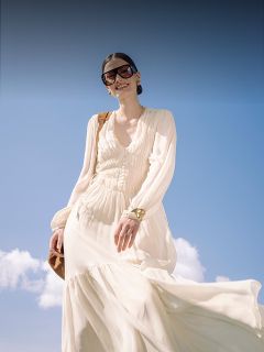 Mujer con un vestido blanco ondeante y grandes gafas de sol sonríe al aire libre frente a un cielo azul con algunas nubes y lleva un bolso marrón.