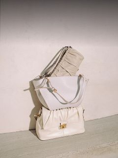 Tres elegantes bolsos de mano en color beige y blanco están apilados y ordenados cuidadosamente frente a una pared clara y un suelo de hormigón a la luz natural del sol.