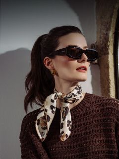 Mujer con gafas de sol oscuras, pendientes dorados, una bufanda con estampado de leopardo y un jersey marrón texturizado posando cerca de una ventana con luz en su rostro.