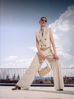 Una mujer con un traje pantalón beige sin mangas y gafas de sol posa al aire libre, con un bolso a juego en la mano, con un paisaje urbano y un cielo azul de fondo.