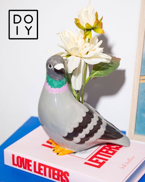 Jarrón de cerámica con forma de paloma con flores blancas sobre un libro de "Cartas de amor", con un logotipo de DOIY en la esquina superior izquierda.