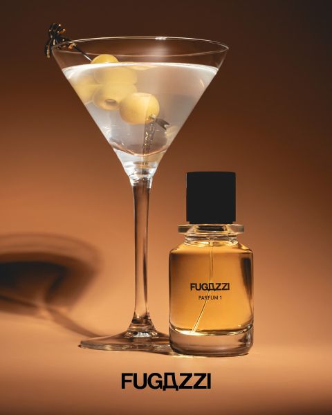 Una copa de Martini con aceitunas junto a un frasco de Fugazzi Parfum I sobre un fondo marrón, debajo el logo de Fugazzi.