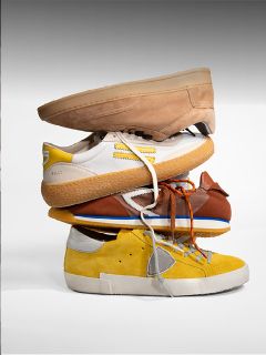 Cuatro zapatillas diferentes en los colores marrón claro, blanco, marrón y amarillo están apiladas una encima de la otra frente a un fondo liso.