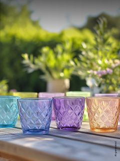 Cuatro vasos de vidrio estructurados y coloridos están sobre una mesa de madera al aire libre, con plantas verdes borrosas y luz solar al fondo.