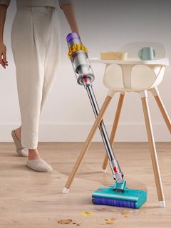 Una persona usa una aspiradora inalámbrica para limpiar un piso de madera cerca de una trona y una mesa pequeña con platos, eliminando las migas.