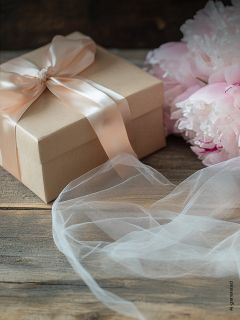 Caja de regalo con lazo rosa pálido junto a peonías rosa pálido y tela blanca sobre una superficie de madera.