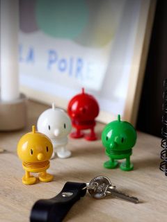 Cuatro figuras de juguete redondas y coloridas están sobre un escritorio junto a llaves y un cuaderno, con una obra de arte enmarcada y borrosa al fondo.