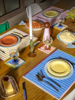 Una mesa de comedor de madera con platos coloridos, manteles individuales acolchados, vasos, cubiertos y velas en una disposición brillante y ecléctica.