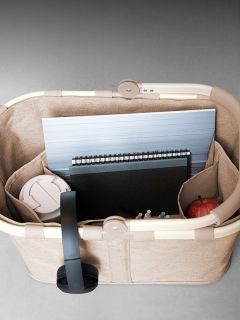 Una cesta beige contiene un portátil, un cuaderno, bolígrafos, un vaso de viaje blanco y una manzana roja, vista desde arriba.